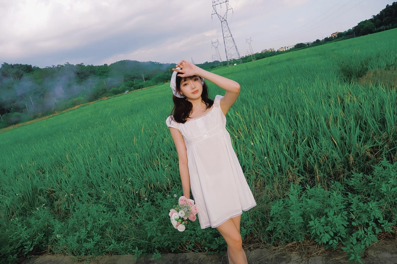 一位穿着白色连衣裙的年轻女子站在绿地上 手里拿着花。