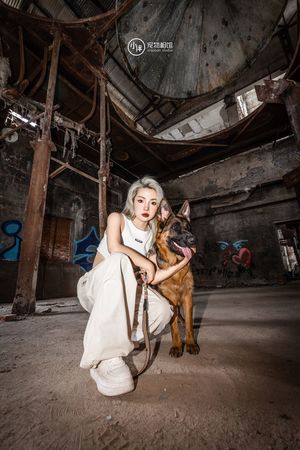 一个穿着白色连衣裙的女人抱着一只狗在一个废弃的仓库或工厂中摆姿势