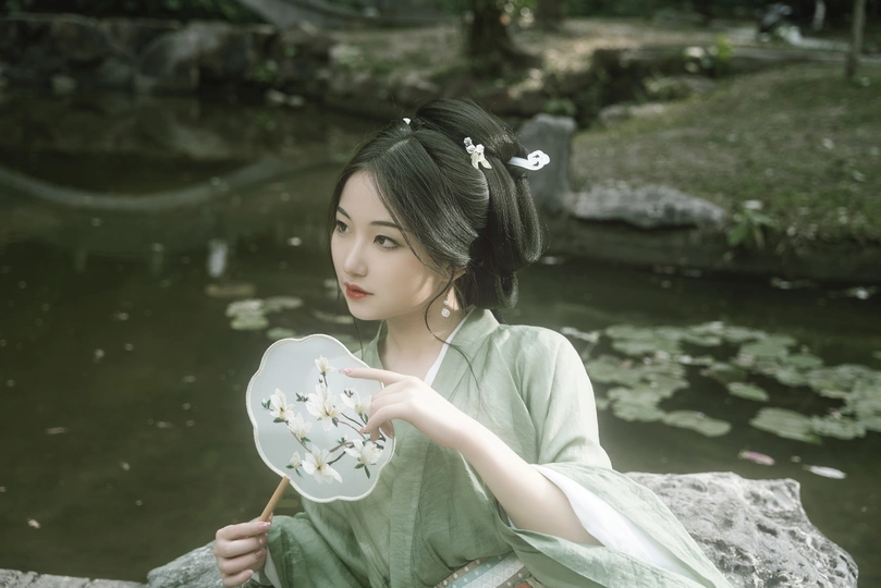 一位穿着绿色连衣裙的年轻女子抱着花坐在池塘前的岩石上