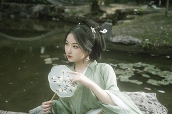 一位穿着绿色连衣裙的年轻女子抱着花坐在池塘前的岩石上