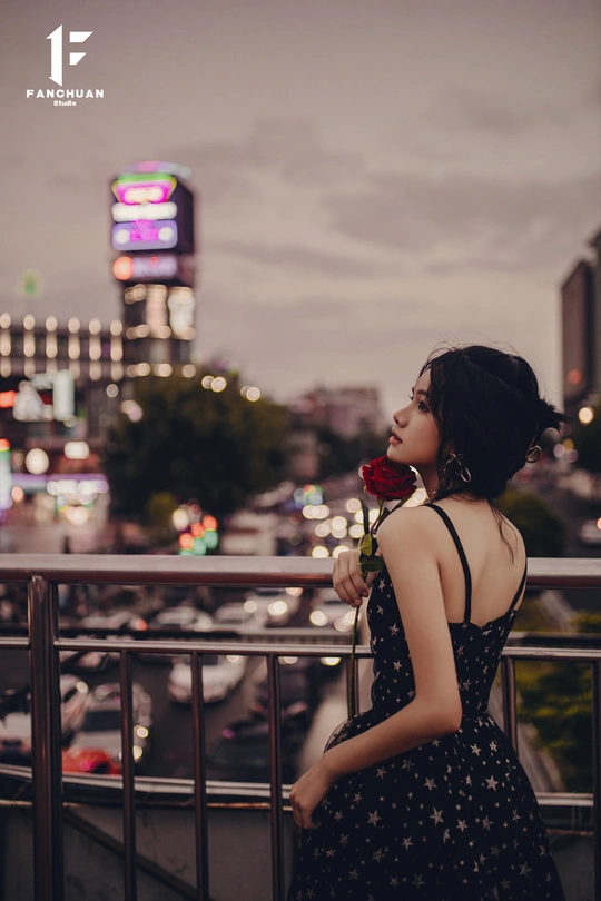 一位穿着连衣裙的女子手拿一朵玫瑰站在阳台上 俯瞰着夜晚的城市。