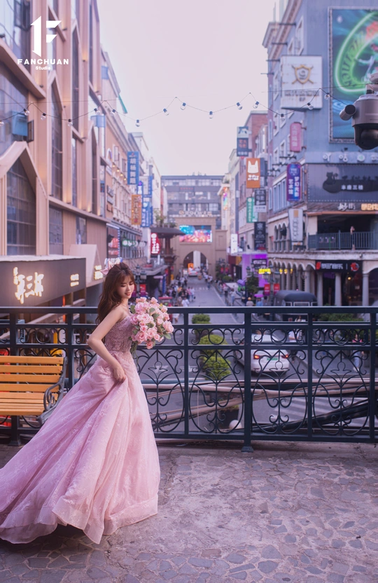 一个穿着粉红色连衣裙 拖尾和一束花的女孩子在城市的一条街道中间站立。