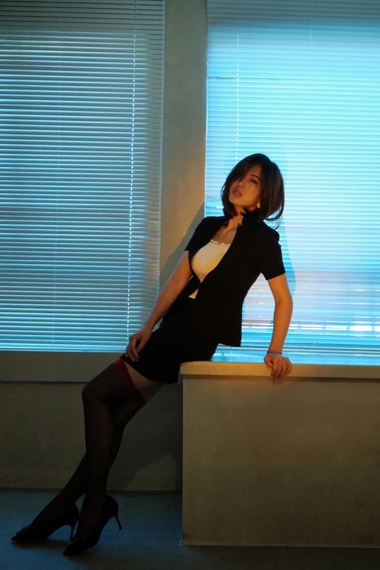 一位穿着黑色连衣裙和高跟鞋的年轻女子坐在办公桌前 面对窗户。