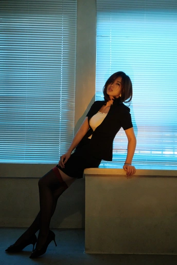 一位穿着黑色连衣裙和高跟鞋的年轻女子坐在办公桌前 面对窗户。