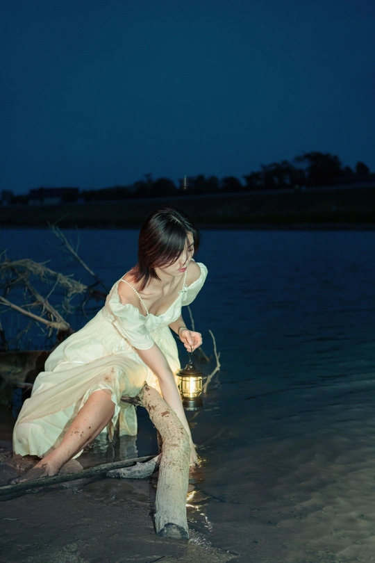 晚上 一个穿着白色连衣裙的年轻女子坐在水中的海滩上 手里拿着一盏亮着的灯笼