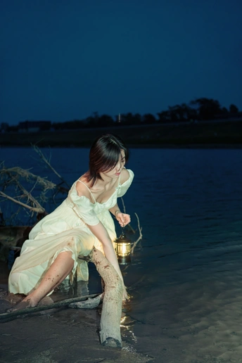 晚上 一个穿着白色连衣裙的年轻女子坐在水中的海滩上 手里拿着一盏亮着的灯笼