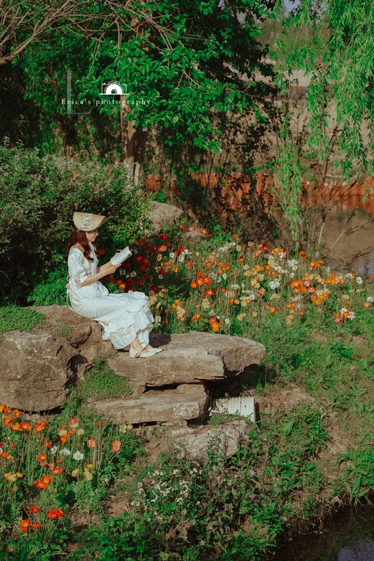 一个穿着白色连衣裙和帽子 坐在溪流边的岩石上阅读书籍的女人