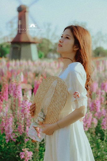 一位穿着白色连衣裙的年轻女子站在一个有粉色花朵和背景风车的小花丛中