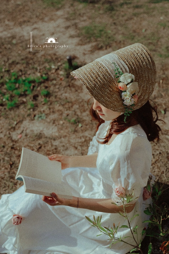 一位穿着白色连衣裙和花朵图案帽子的小女孩在户外看书。
