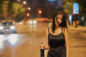 一位穿着黑色连衣裙的年轻女子手拿一瓶饮料和一只手提箱站在城市街道的 sidewalk 之夜