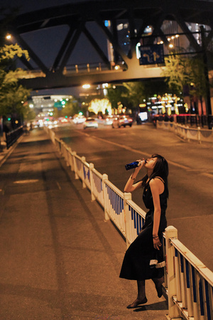 一个穿着黑色连衣裙的女人站在一座桥上 夜晚时分 她正在俯视着公路 手里拿着一瓶水 靠在围墙上。