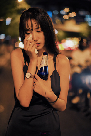 一个穿着黑色连衣裙的年轻女子手持一瓶饮料 站在夜晚的街道上 正在用手机通话。