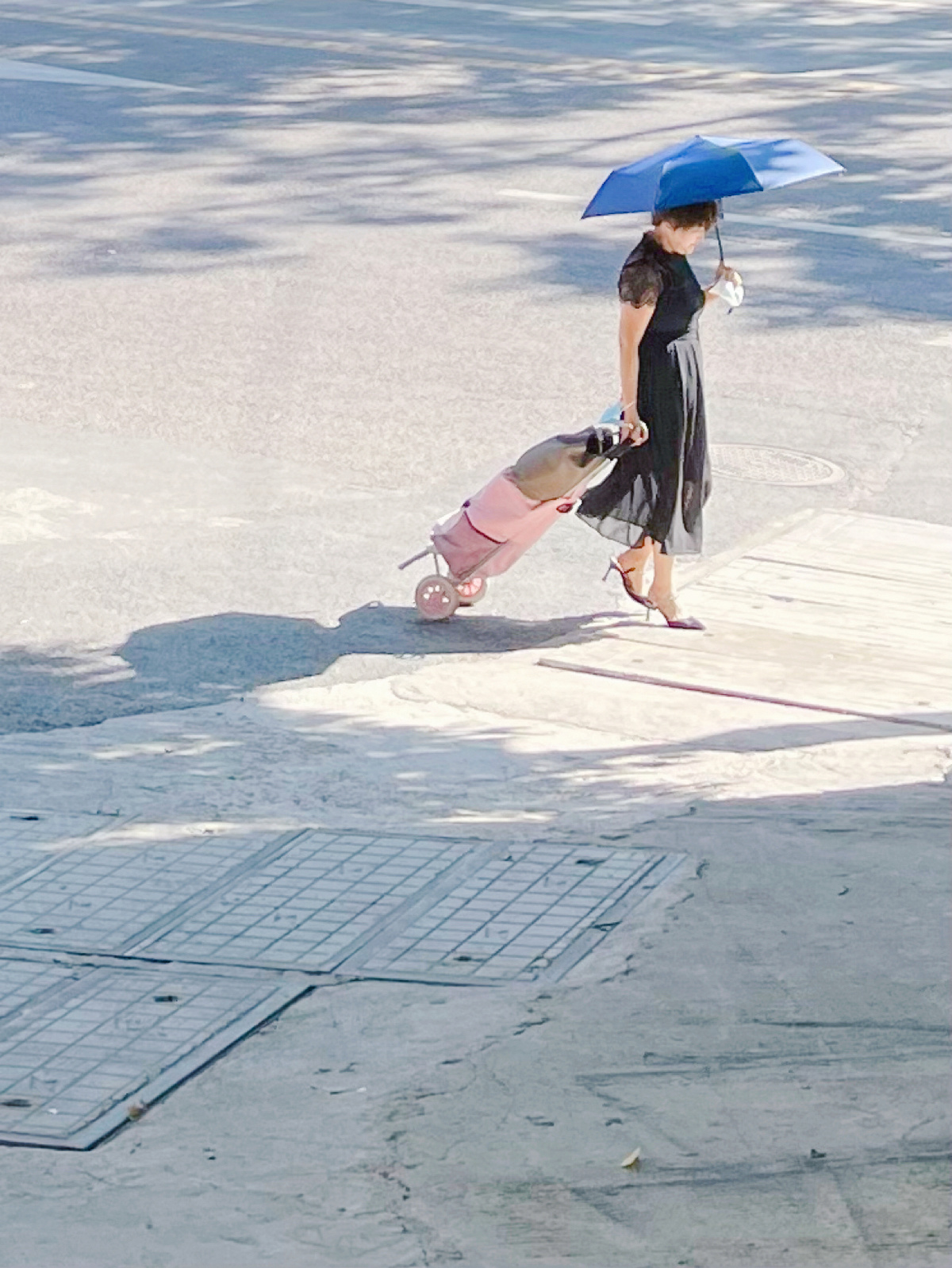 一位穿着黑色连衣裙的年轻女子拿着一把蓝色雨伞和一只粉色手提箱 而一位穿着黑色连衣裙的小女孩正在街上走